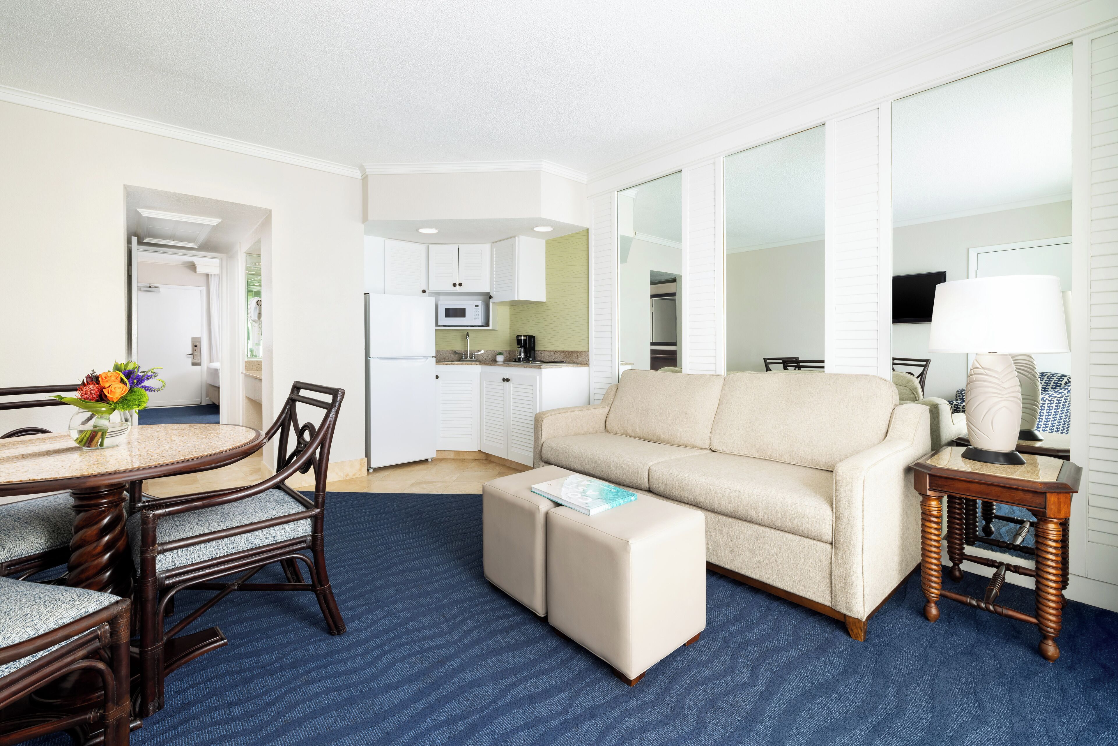 A modern living room with a beige sofa, a round dining table with chairs, a small ottoman, a side table with a lamp, and a kitchen area in the background.