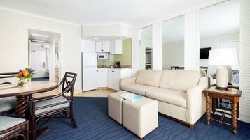 A modern living room with a beige sofa, a round dining table with chairs, a small ottoman, a side table with a lamp, and a kitchen area in the background.
