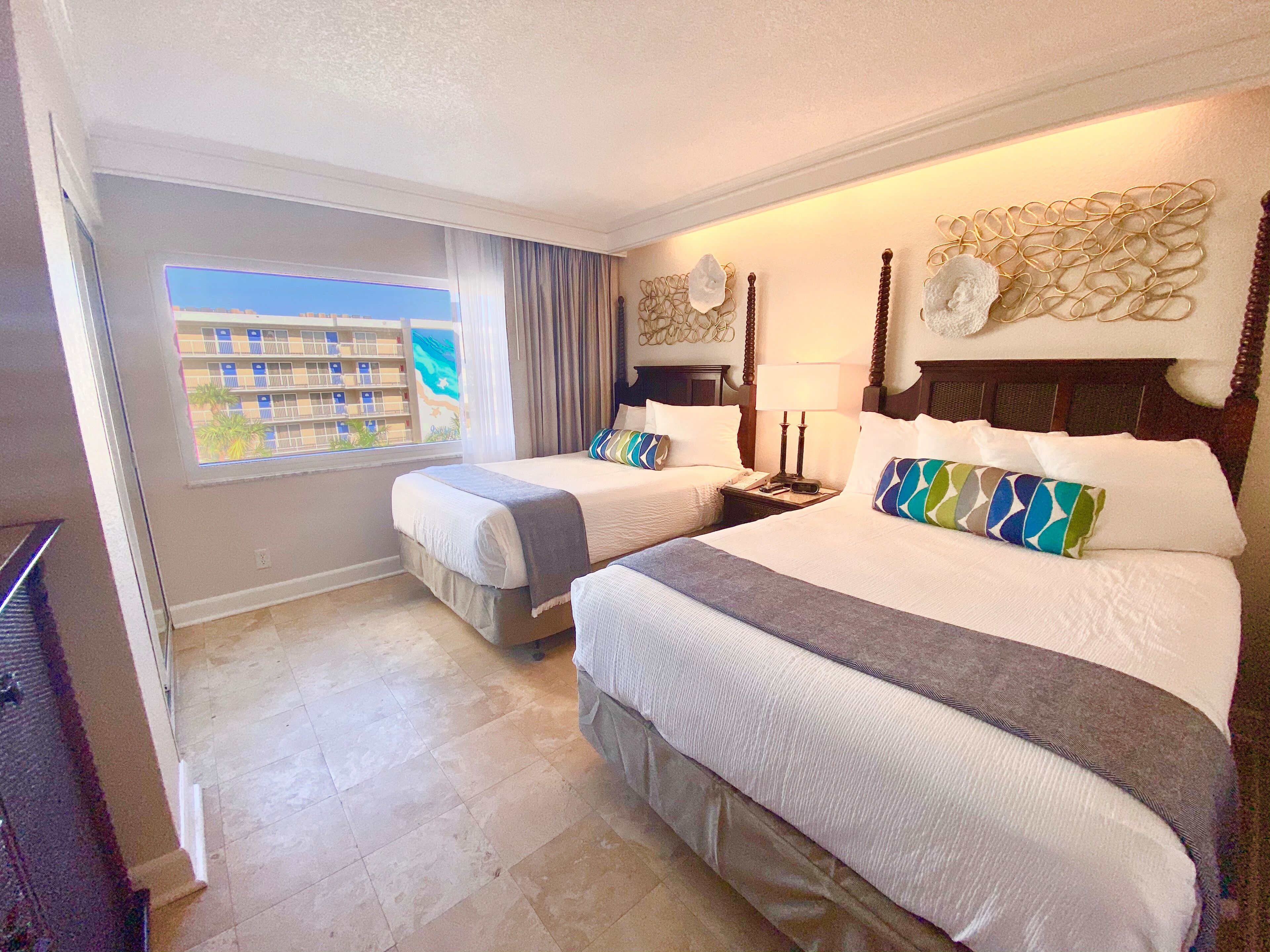A hotel room with two beds, a window with a view of a building, a lamp, and a decorative wall piece.