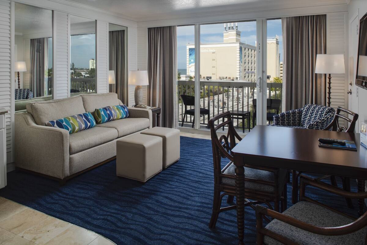 A hotel room with a large window offering a city view, a sofa with patterned cushions, a dining table with chairs, and a blue textured rug.