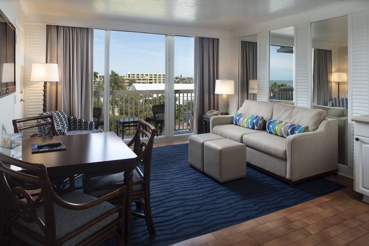 A hotel room with a dining table, chairs, a sofa, and a balcony with a view.