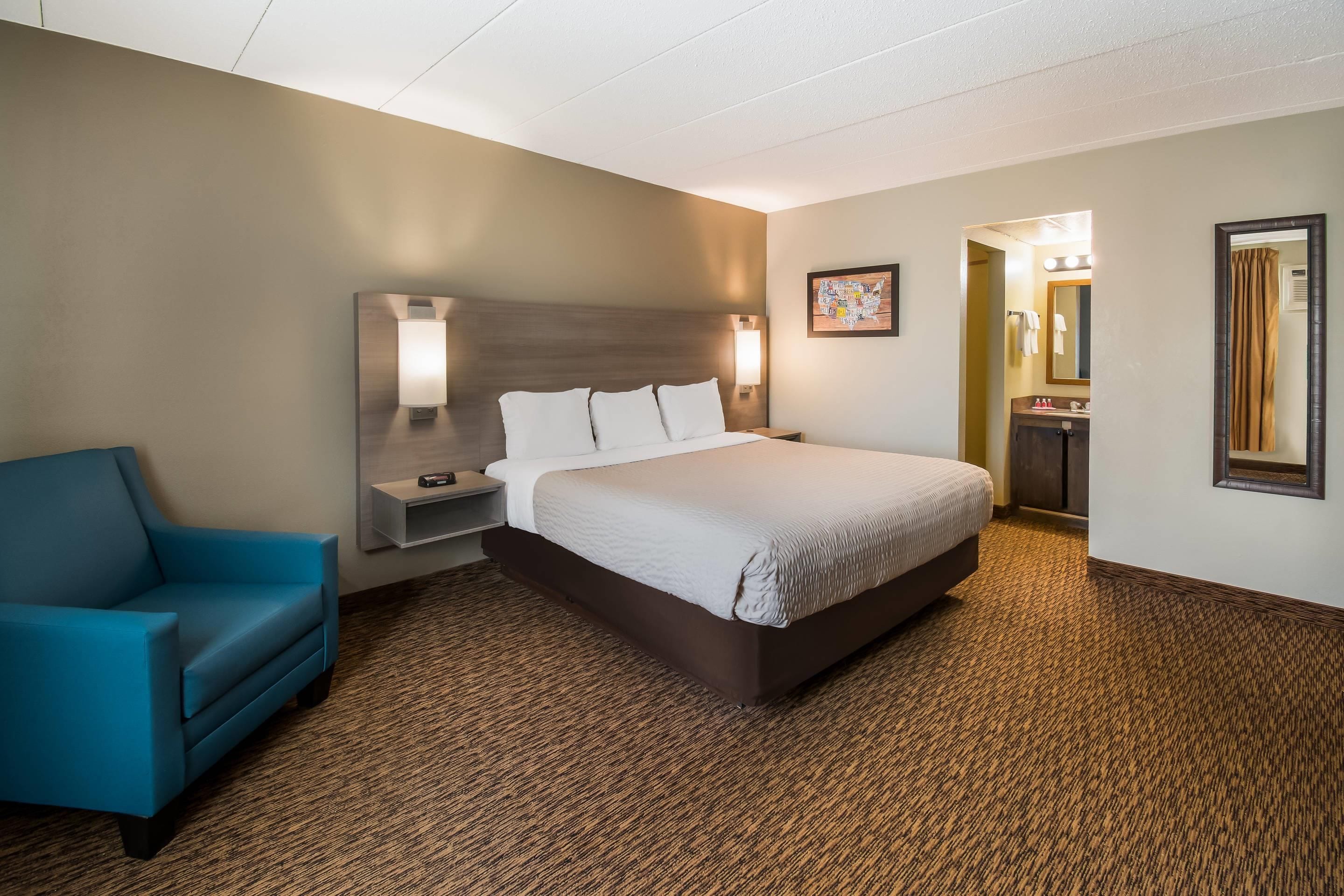 Habitación de hotel con cama, mesita de noche, una silla azul, lámpara fijada a la pared y un baño visible a través de una puerta abierta.