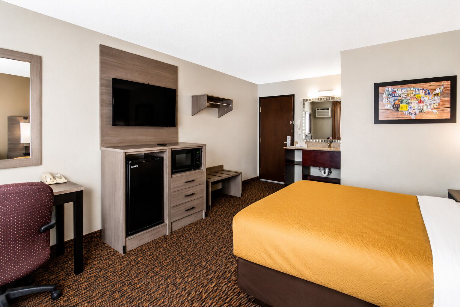 A hotel room with a bed, a desk with a microwave, a chair, a wardrobe, a television, and a bathroom with a sink and mirror.