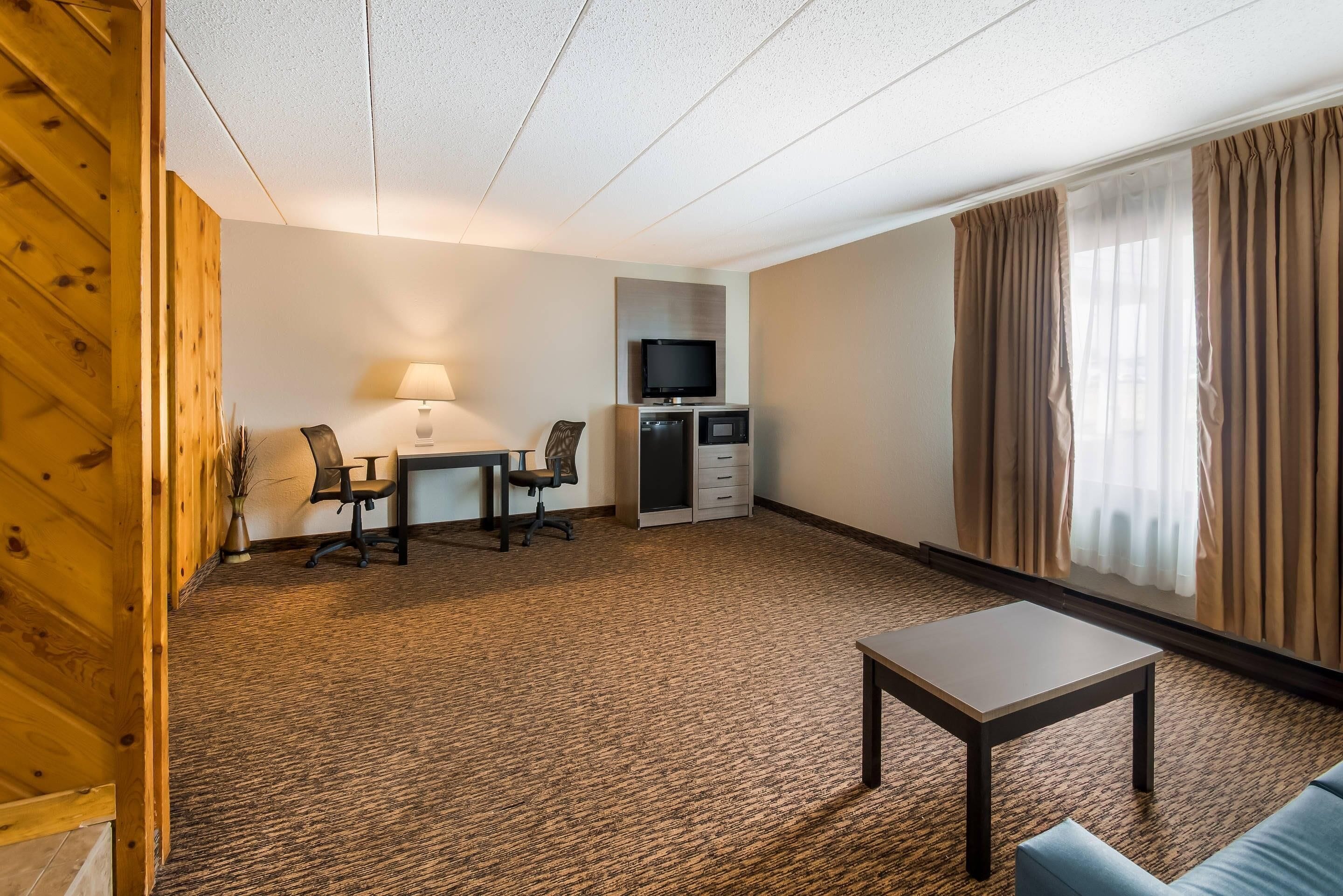 Ein Hotelzimmer mit Schreibtisch, Stühlen, Fernseher, einer kleinen Küchenzeile, einer Couch und einem Fenster mit Vorhängen.