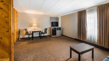 Ein Hotelzimmer mit Schreibtisch, Stühlen, Fernseher, einer kleinen Küchenzeile, einer Couch und einem Fenster mit Vorhängen.