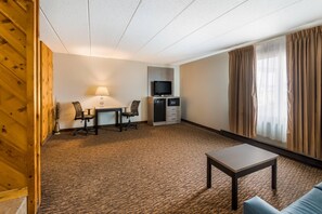 A hotel room with a desk, chairs, a television, a small kitchenette, a sofa, and a window with curtains.