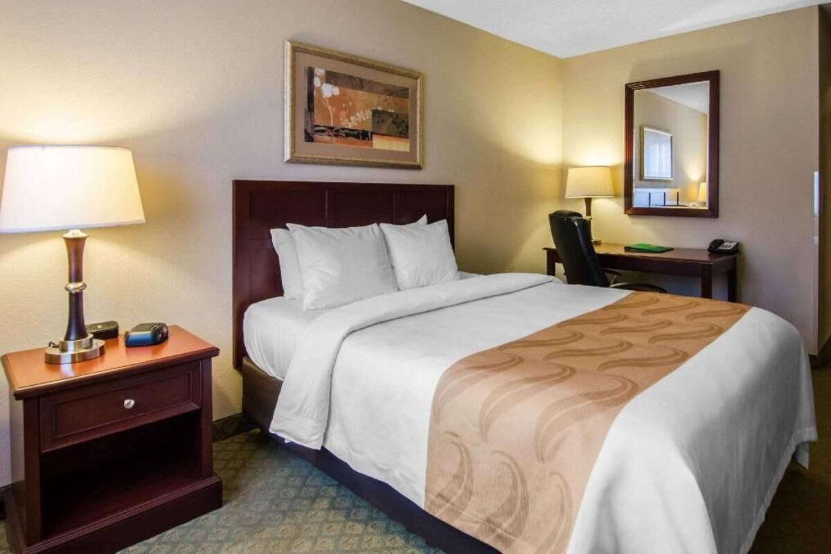 A hotel room with a bed, a nightstand with a lamp, a desk with a chair, and a mirror.