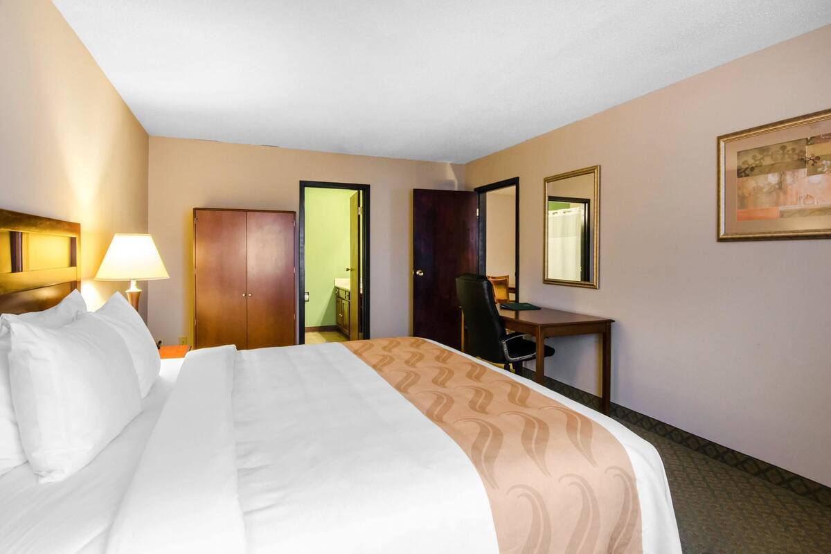 A hotel room with a bed, a desk, a chair, a lamp, and a door.