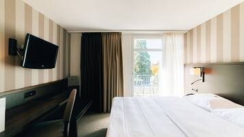 Quarto de hotel com cama, escrivaninha com cadeira, televisão e janela com cortinas.