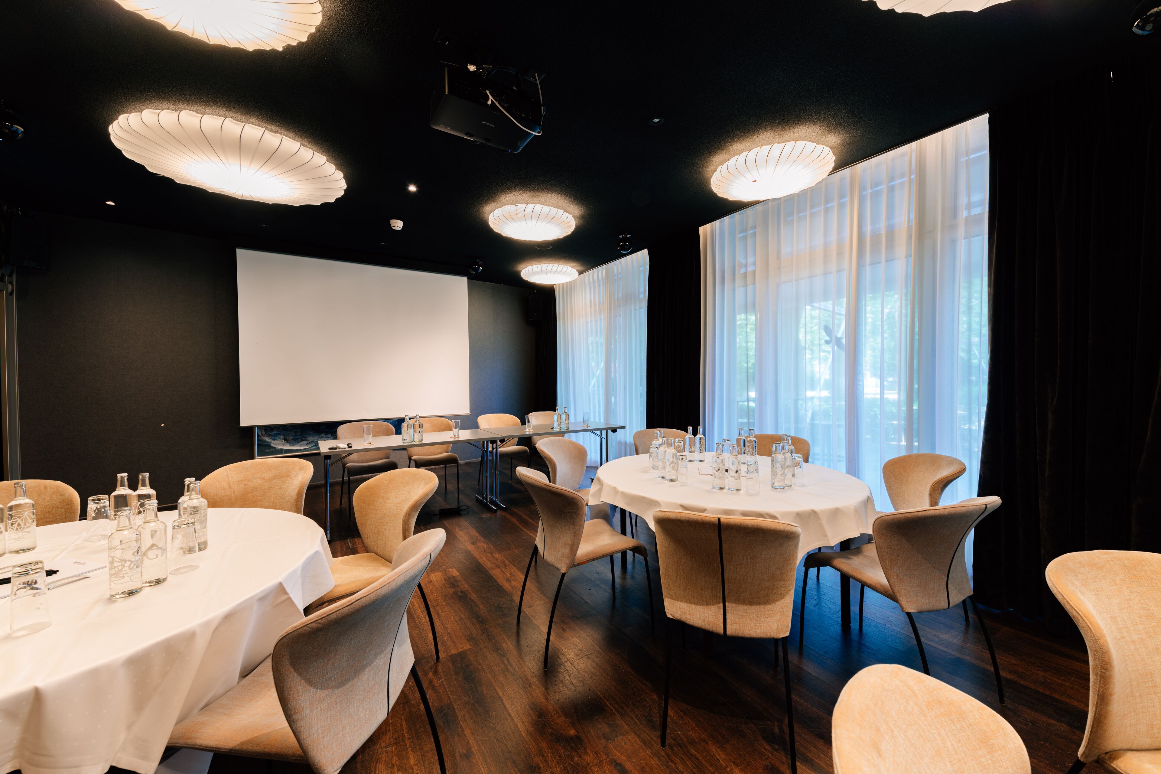 Ein Konferenzraum mit runden Tischen, Stühlen, einer Projektionsleinwand und großen Fenstern mit transparenten Vorhängen.