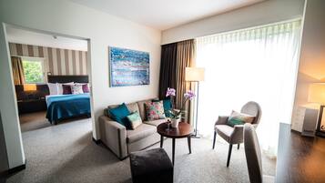 Habitación de hotel con cama, sofá, mesa de centro y una silla. Se observa un cuadro en la pared y una ventana con cortinas.