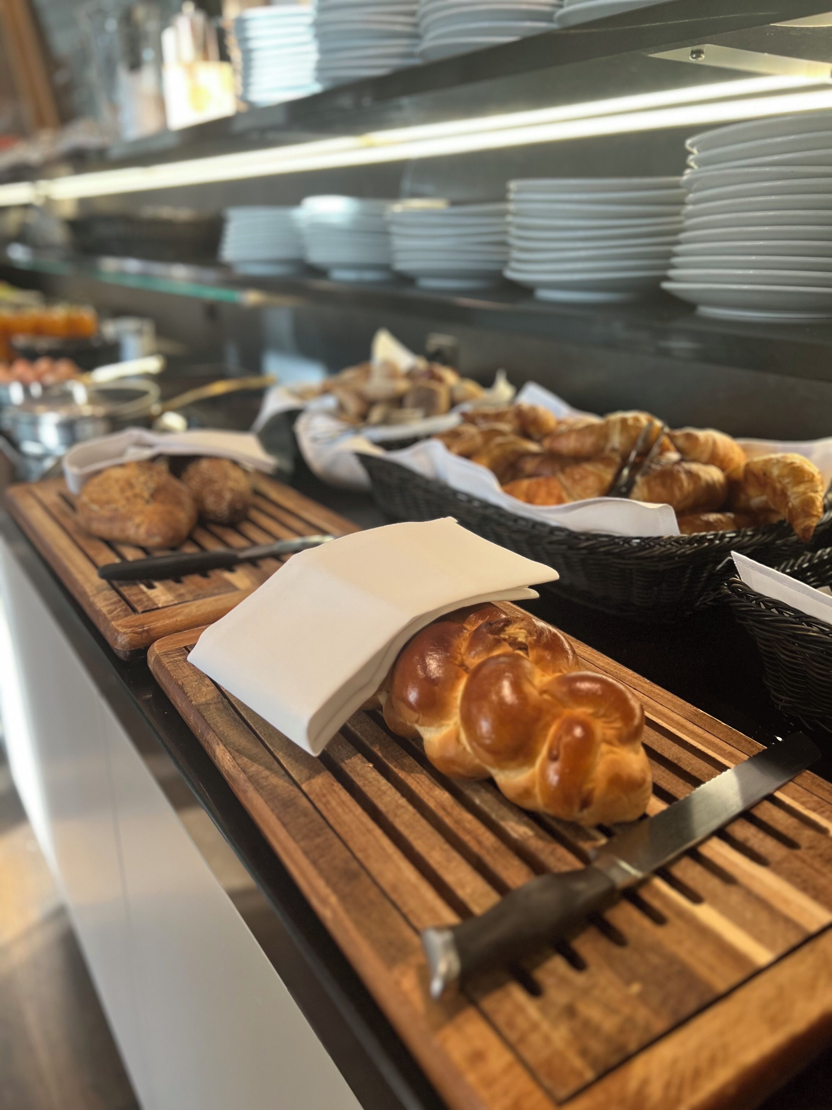 Ein Frühstücksbuffet mit verschiedenen Backwaren und Brot, darunter Croissants und einem Zopf, auf Holzbrettern.