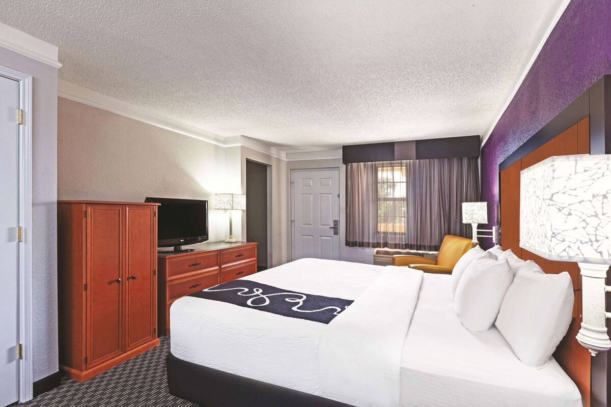 A hotel room with a bed, a television, a desk with a lamp, a chair, and a window with curtains.
