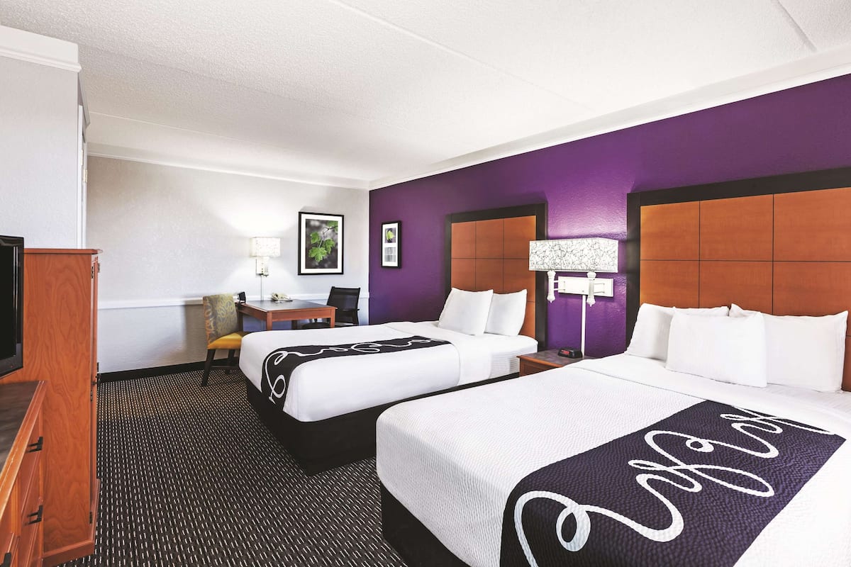 A hotel room with two beds, a desk, a chair, a TV, and a lamp.