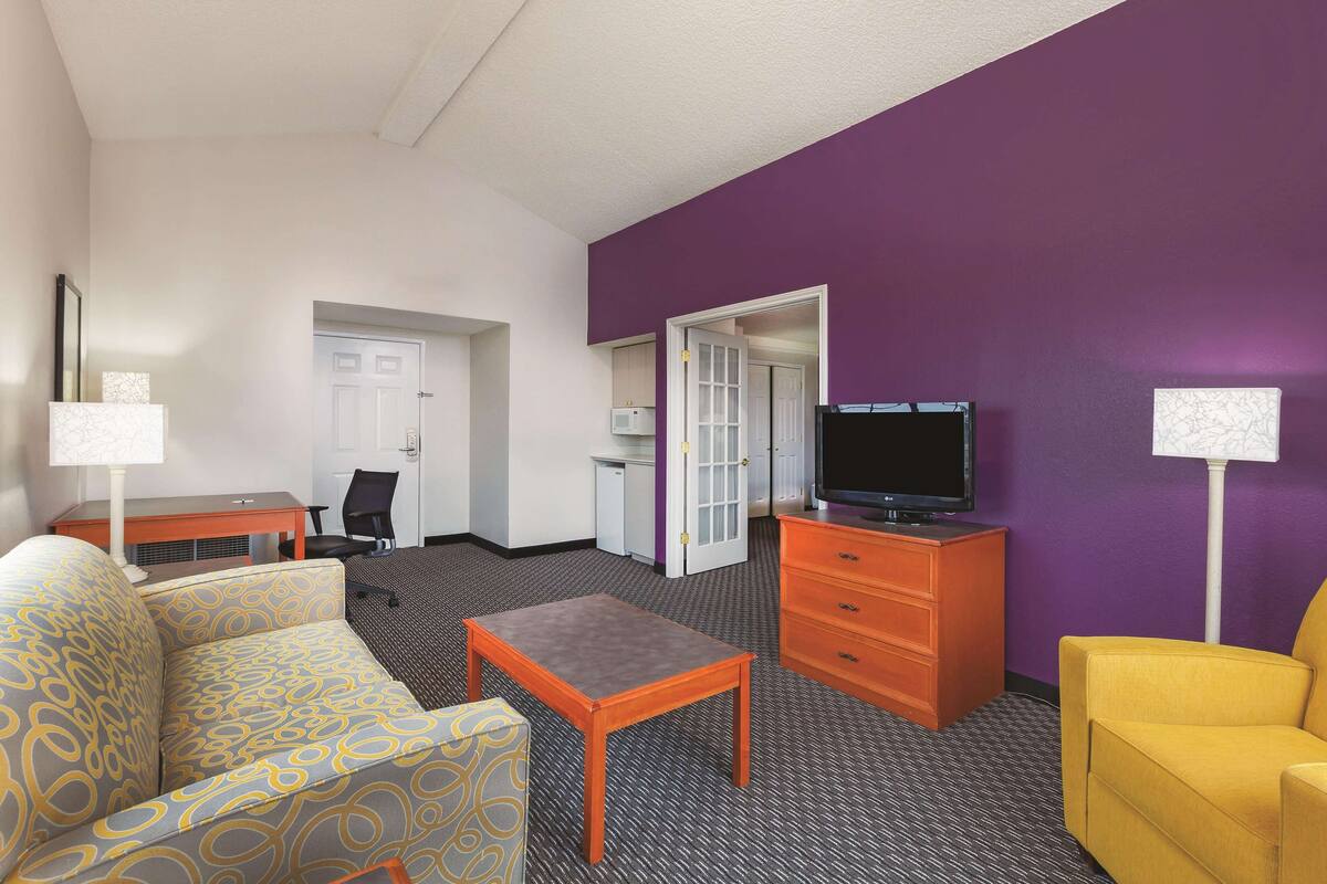 A hotel room with a sofa, a small table, a TV on a wooden stand, and a yellow armchair.