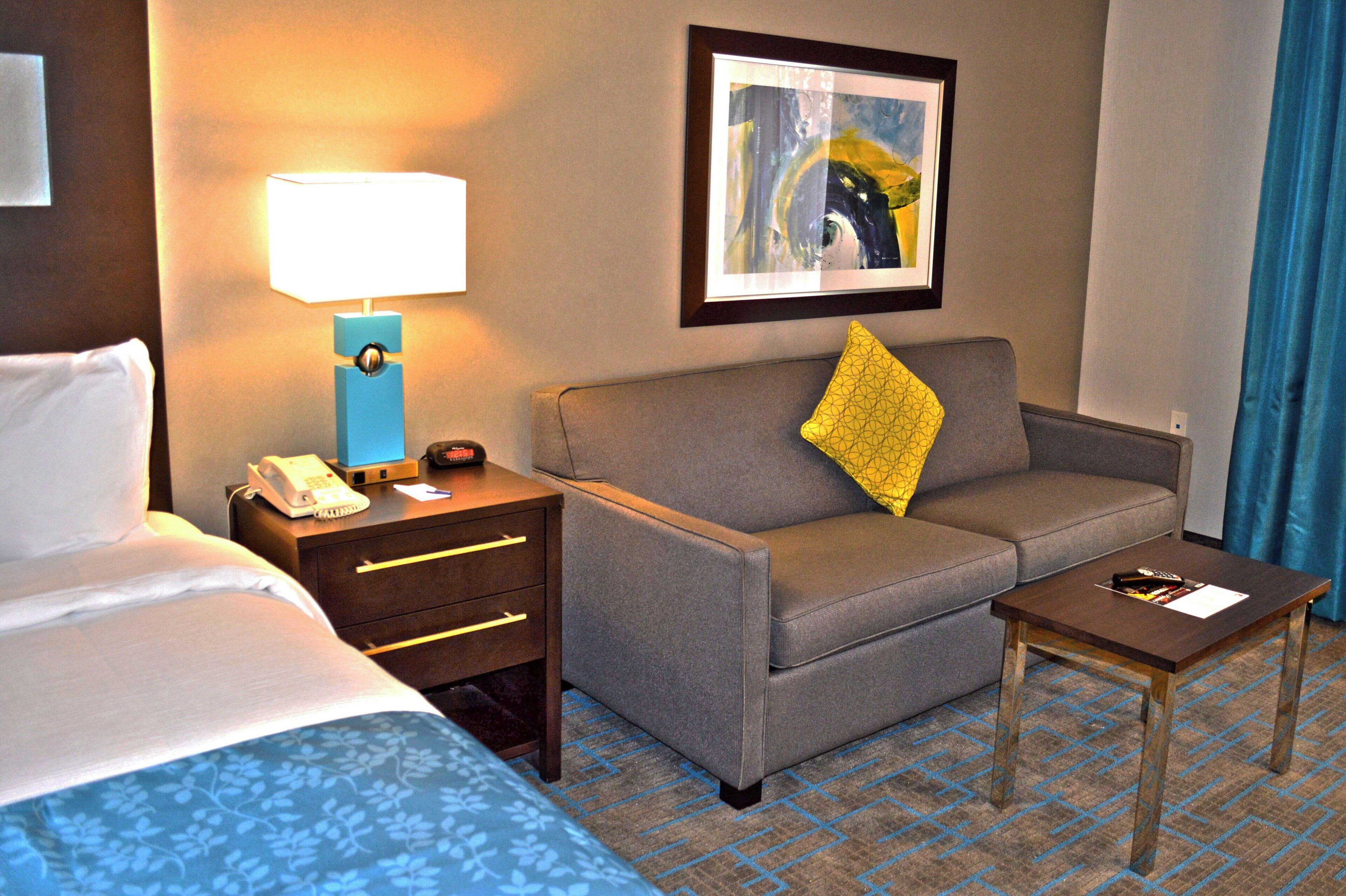 A hotel room with a bed, a desk with a lamp, a sofa with a pillow, and a small table with a remote control.