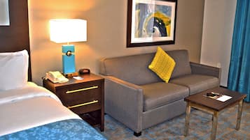 A hotel room with a bed, a desk with a lamp, a sofa with a pillow, and a small table with a remote control.