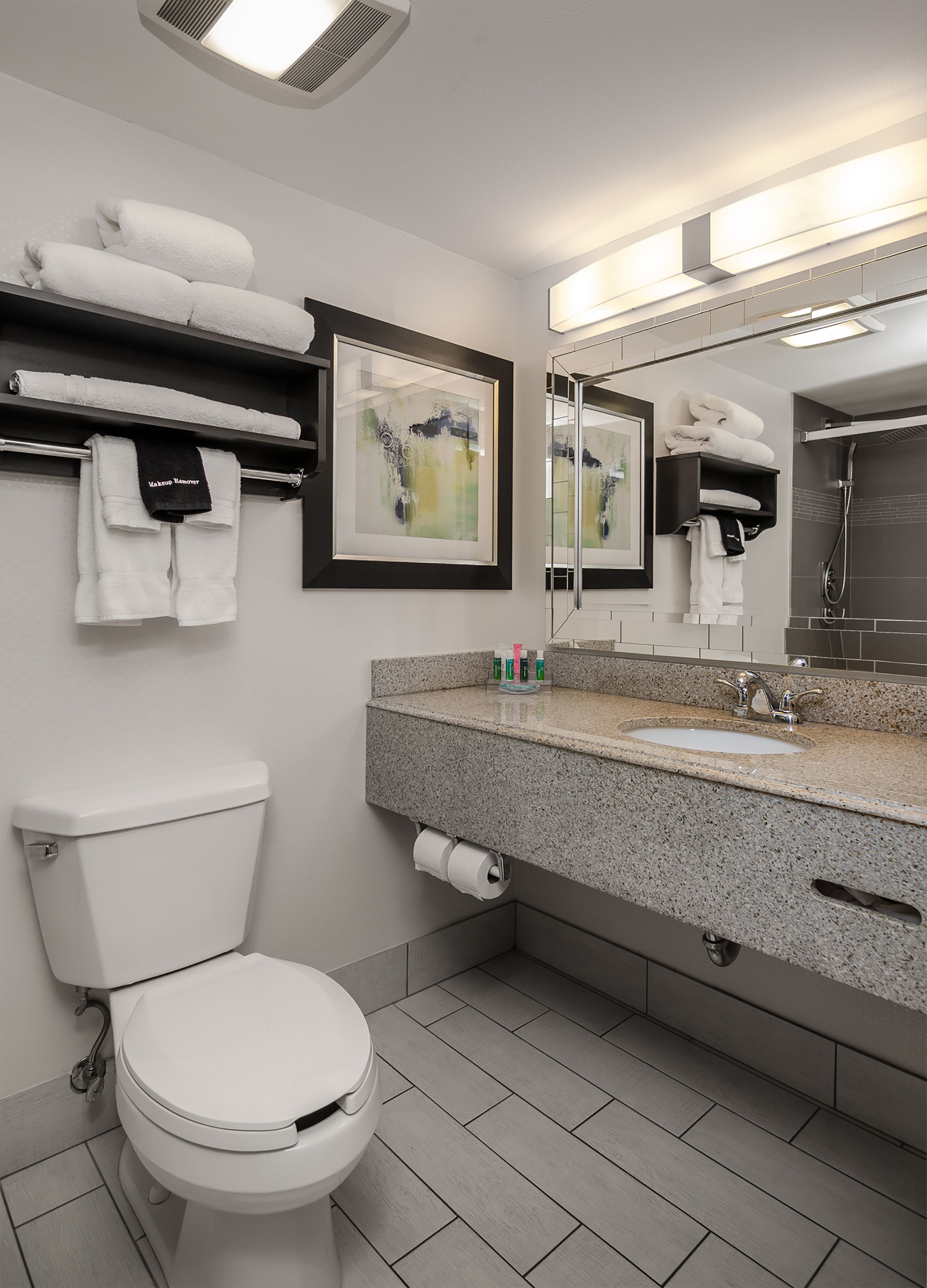 A modern bathroom with a toilet, sink, and shower. There are towels on a shelf and a framed abstract painting on the wall.