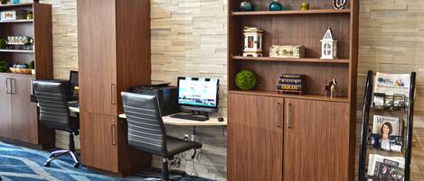 A hotel room with a wooden desk, two office chairs, a computer, a bookshelf with decorative items, and a magazine rack.
