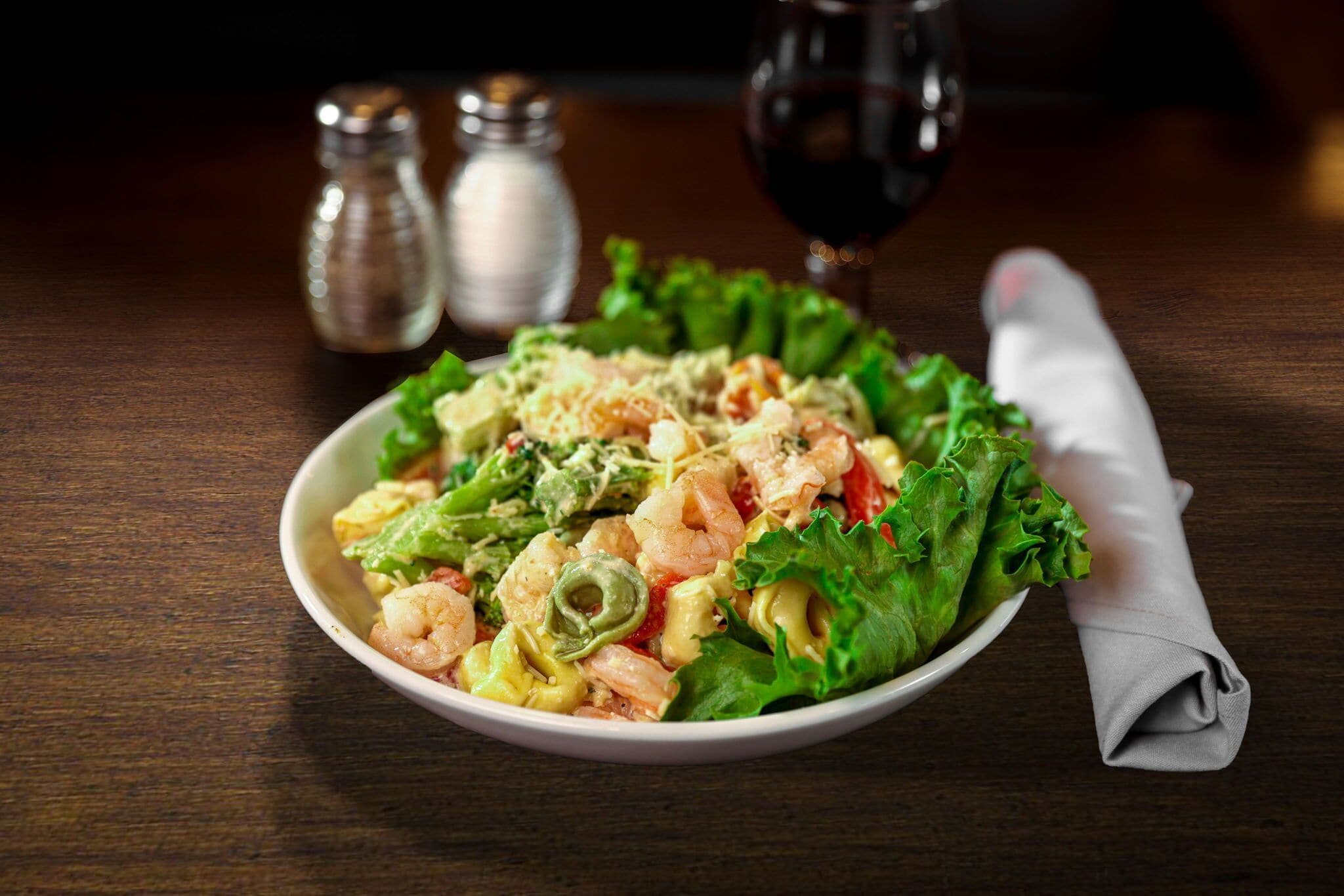 Un tazón de ensalada de pasta con camarones, una servilleta y saleros y pimenteros sobre una mesa de madera.