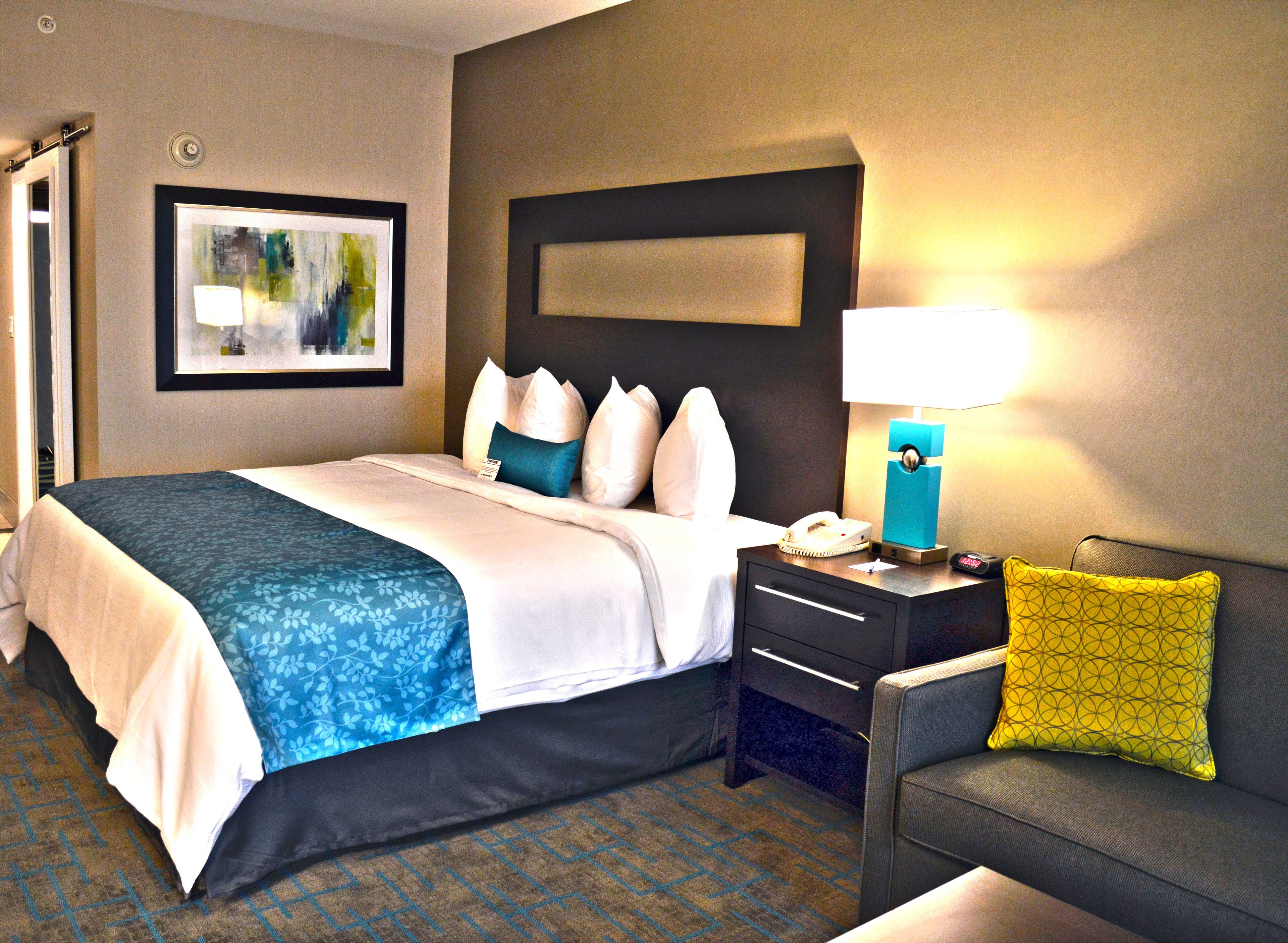 A hotel room with a bed, a nightstand, a lamp, a chair, and a painting on the wall.