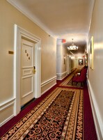 Um corredor de hotel com tapete vermelho, portas brancas e um candelabro.