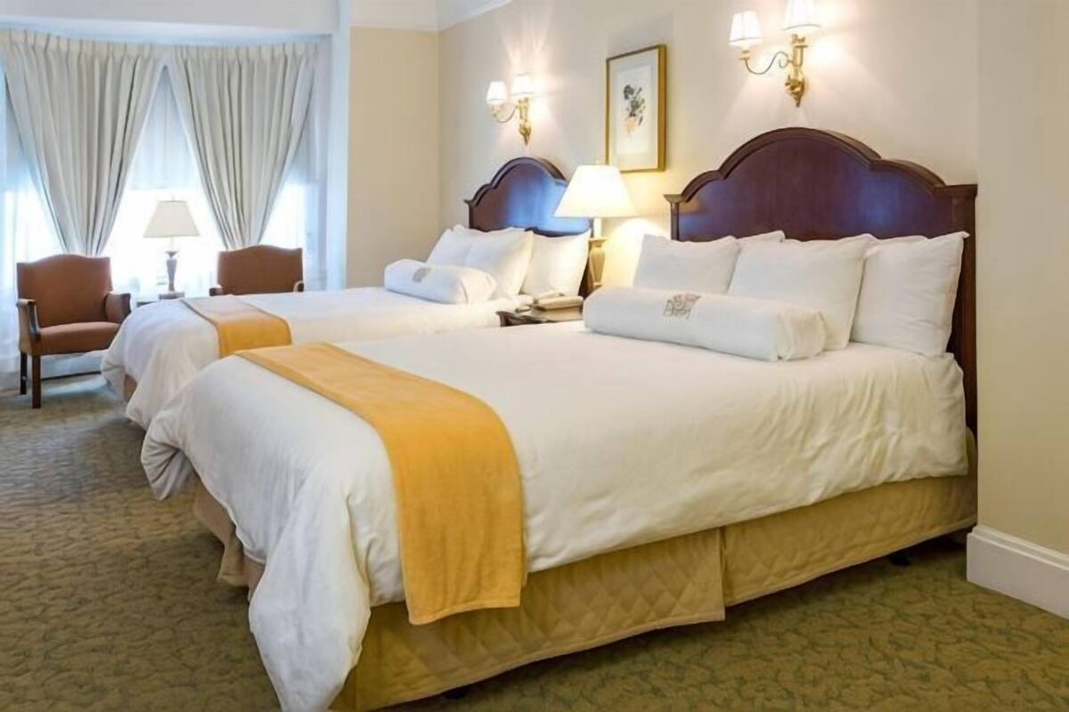 Ein Hotelzimmer mit zwei Betten, einem Holzkopfstück, Nachttischlampen, einem Sessel und einem Teppichboden.