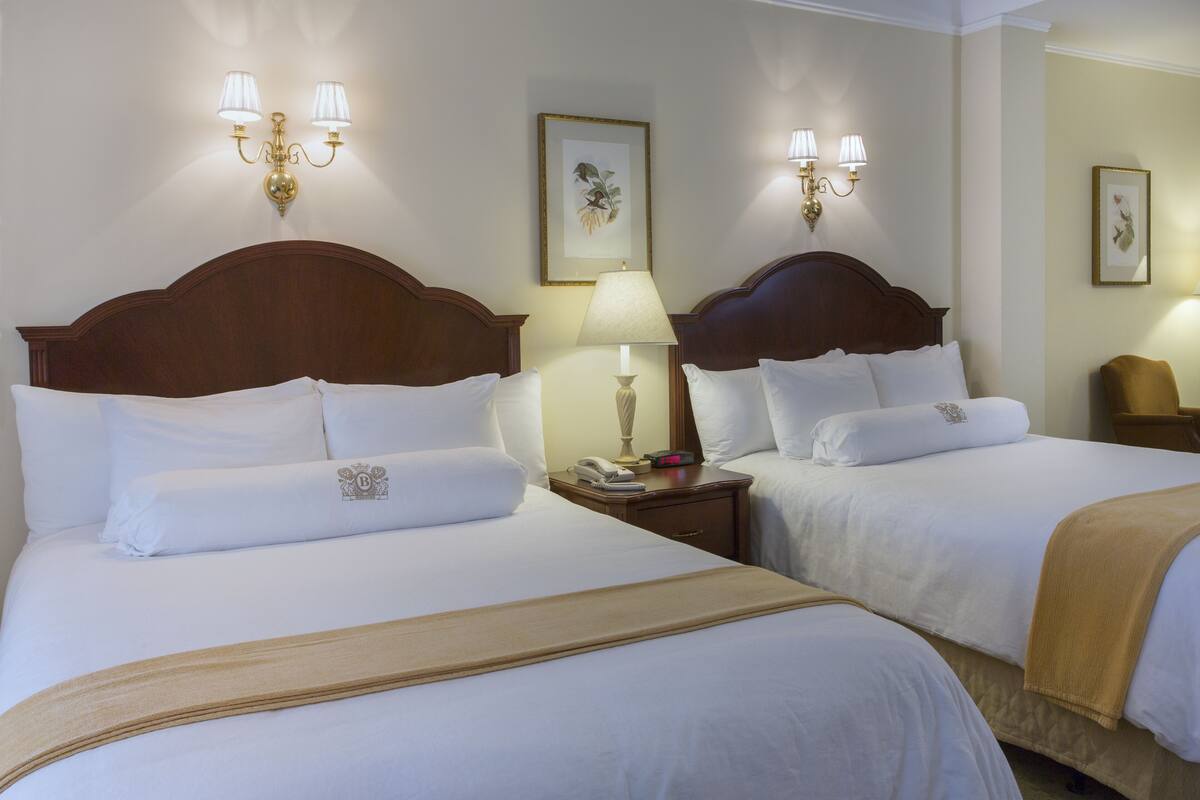 Ein Hotelzimmer mit zwei Betten, jeweils mit weißer Bettwäsche und einem hölzernen Kopfteil. An den Wänden sind Wandlampen und ein gerahmtes Bild angebracht.