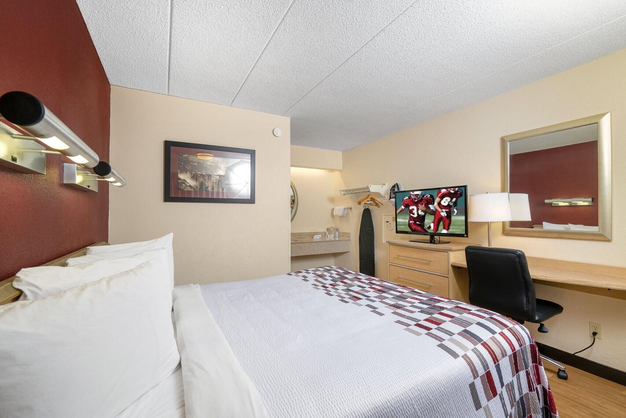 Habitación de hotel con cama, escritorio, silla, televisor, espejo y un cuadro en la pared.