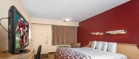 Quarto de hotel com cama, escrivaninha com televisão, cadeira, luminária de mesa e luminária na parede.
