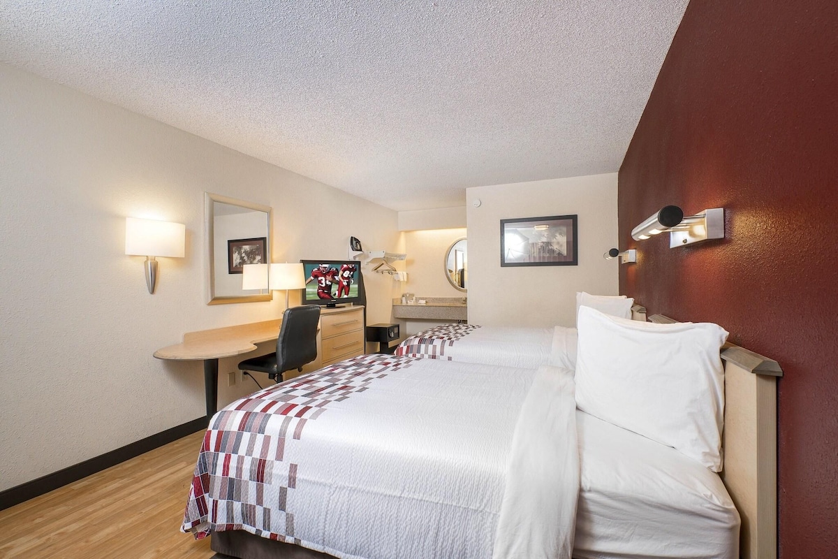 A hotel room with two beds, a desk, a chair, a mirror, and wall-mounted lights.