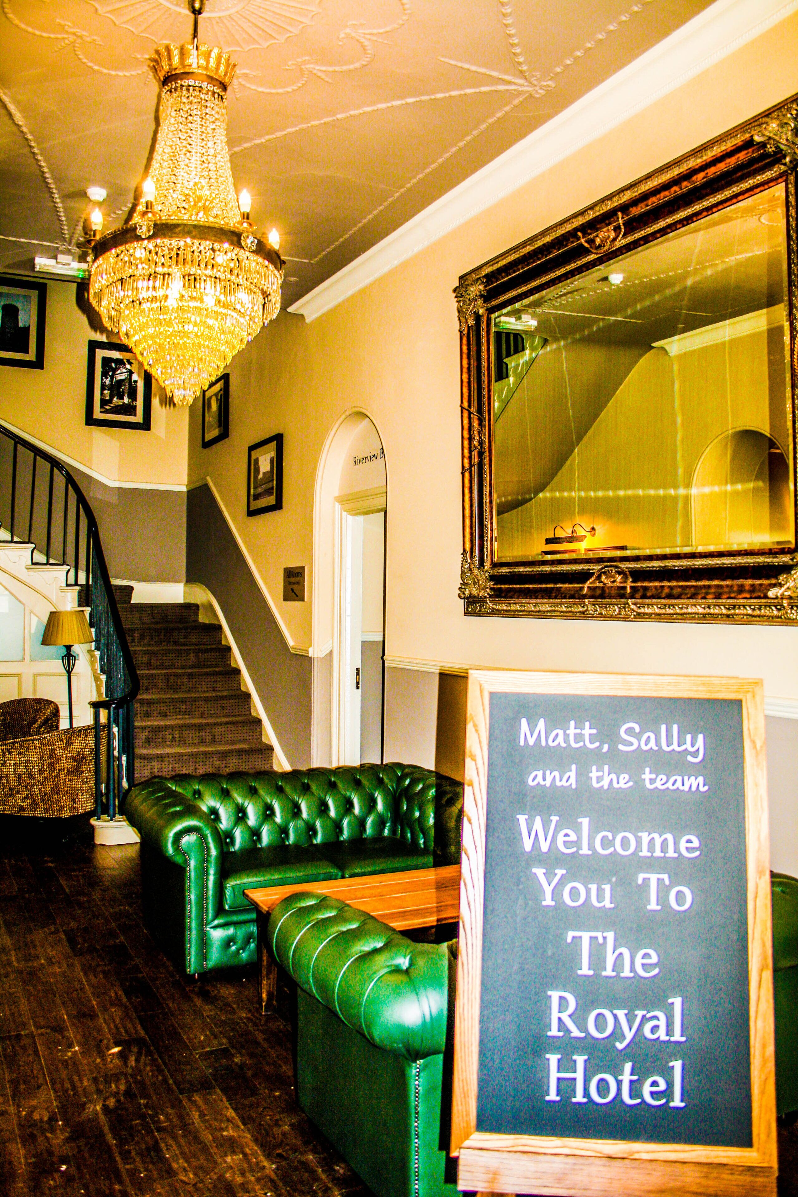 A grand lobby with a chandelier, a green leather sofa, a wooden coffee table, and a sign welcoming guests to the Royal Hotel.