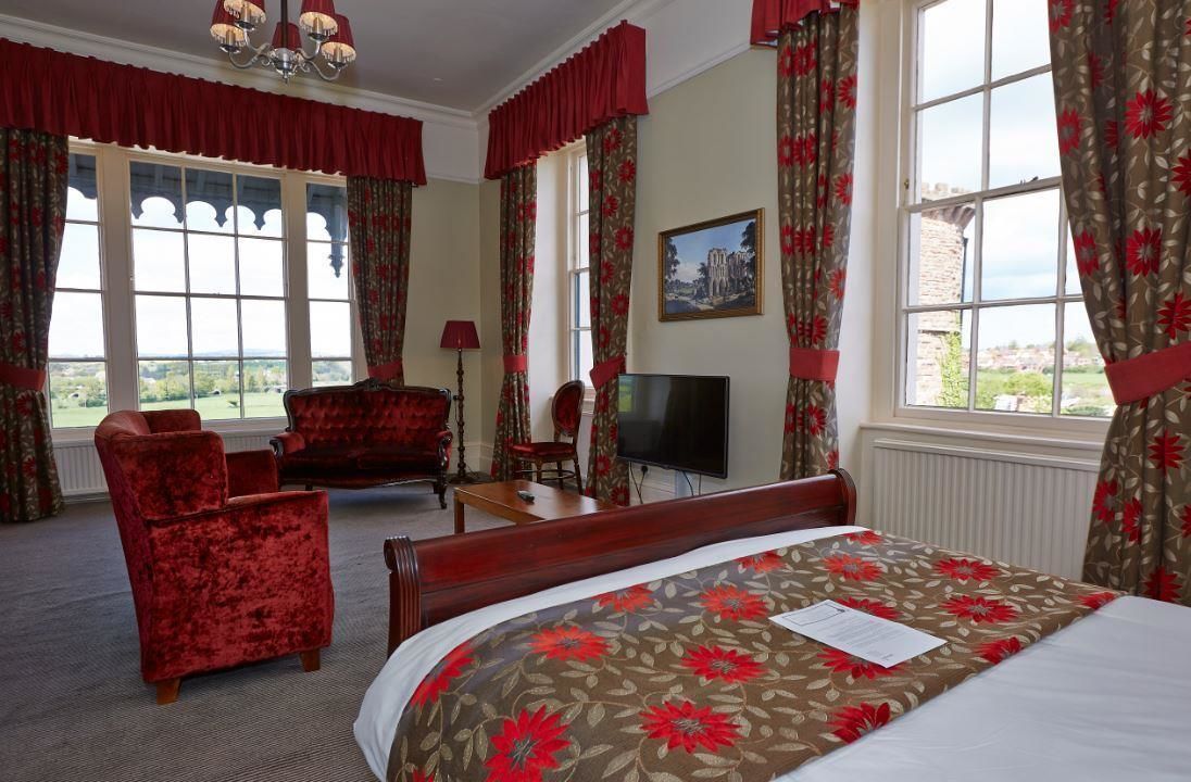 Habitación de hotel con una cama grande, dos sillones rojos, un televisor y vistas al campo.