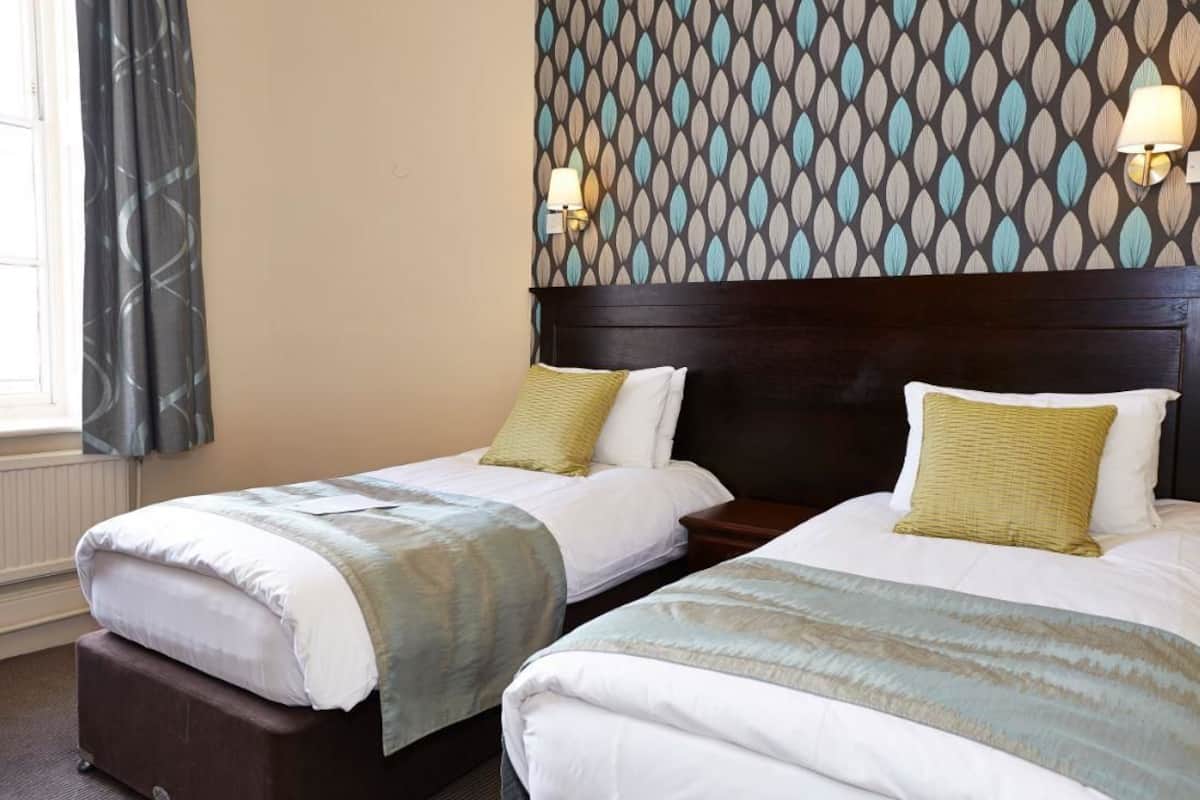 Hotelkamer met twee enkele bedden, elk voorzien van een witte dekbedovertrek en een groen kussen. De kamer heeft een donker houten hoofdbord, behang met een patroon en twee wandlampen.