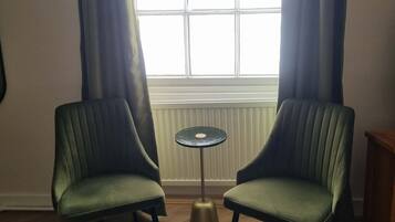 Une pièce avec deux fauteuils verts, une petite table ronde et une fenêtre avec des rideaux.