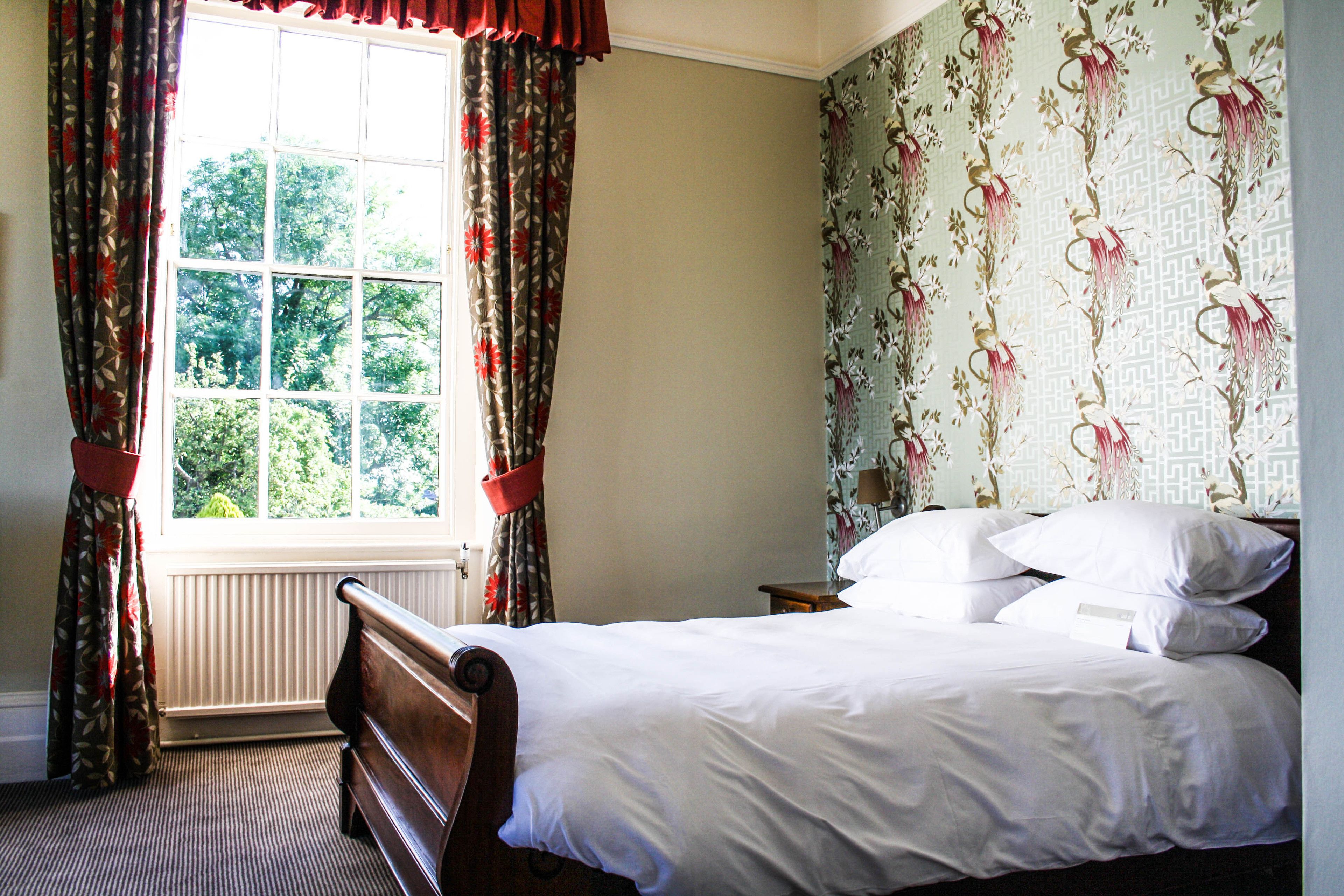 Een slaapkamer met een groot raam, een bed met wit beddengoed en bloemenbehang.
