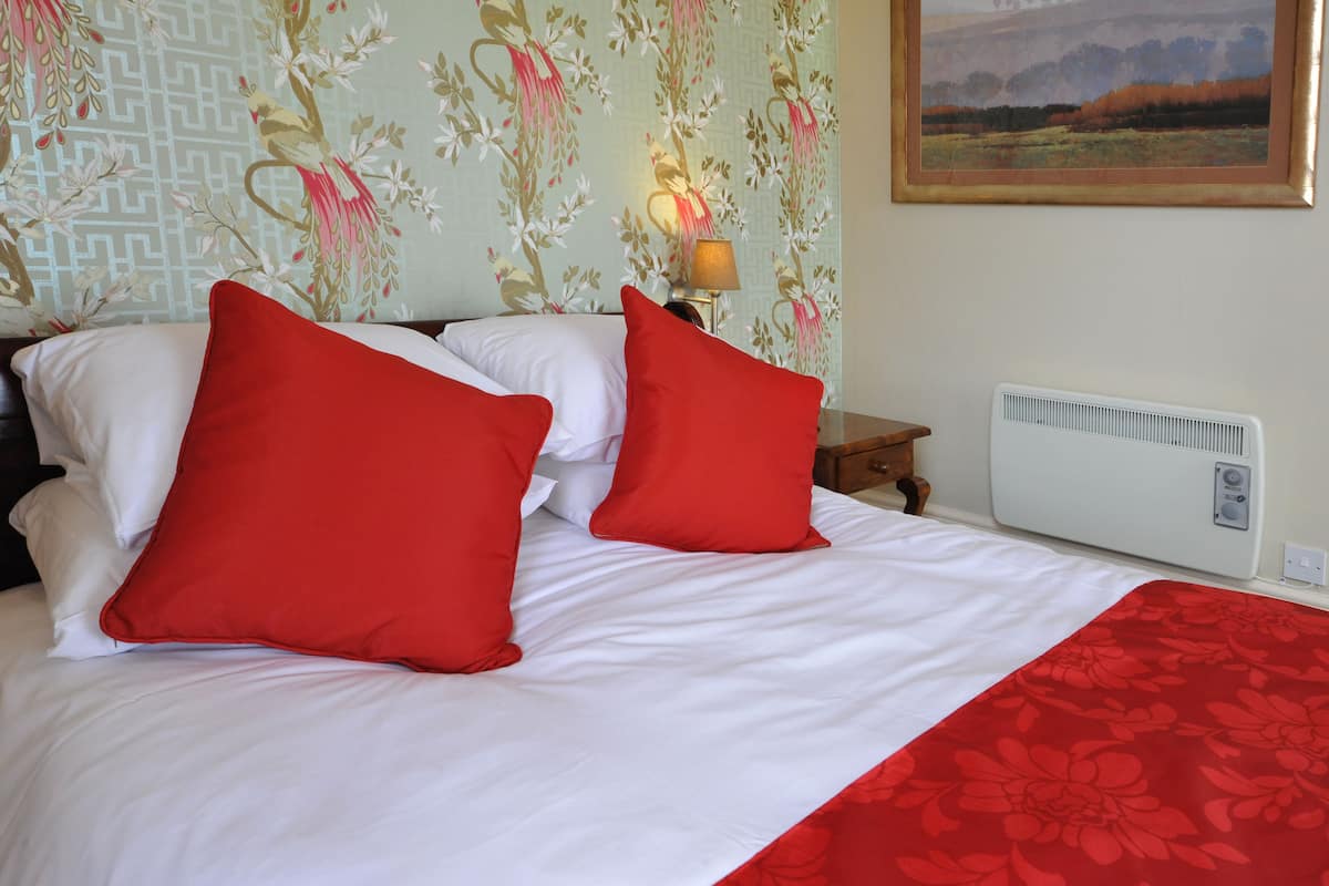 Een net opgemaakt bed met wit en rood beddengoed, een nachtkastje met een lamp, en een muur met bloemenbehang en een lijst met een landschapsafbeelding.
