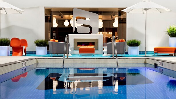 A modern indoor pool area with a clear pool, orange and gray seating, and a central fireplace. - Aloft by Marriott Calgary University (Calgary)