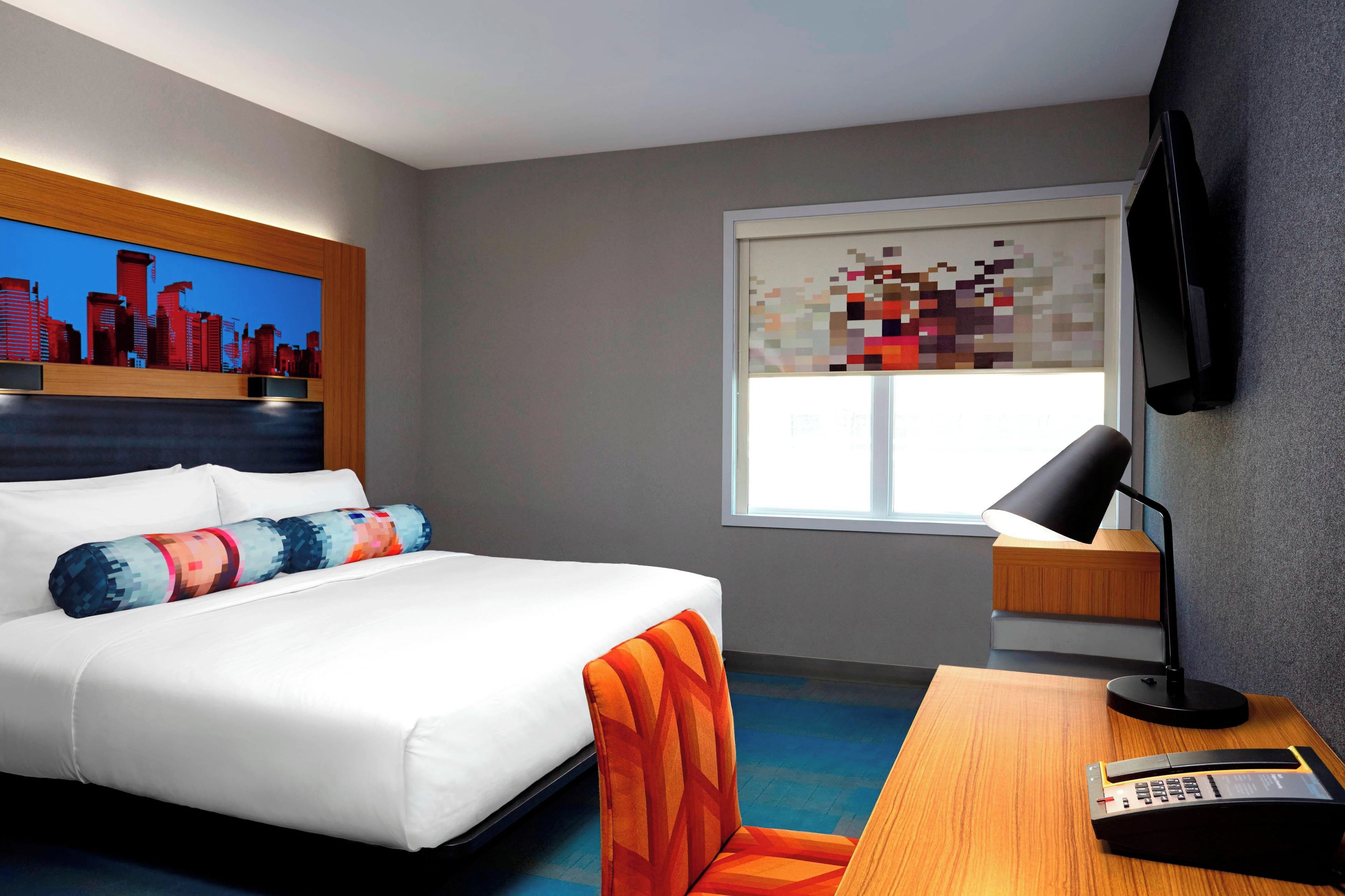 A hotel room with a bed, a desk with a lamp, a cityscape painting, and a pixelated artwork on the window.