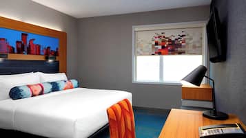 A hotel room with a bed, a desk with a lamp, a cityscape painting, and a pixelated artwork on the window.
