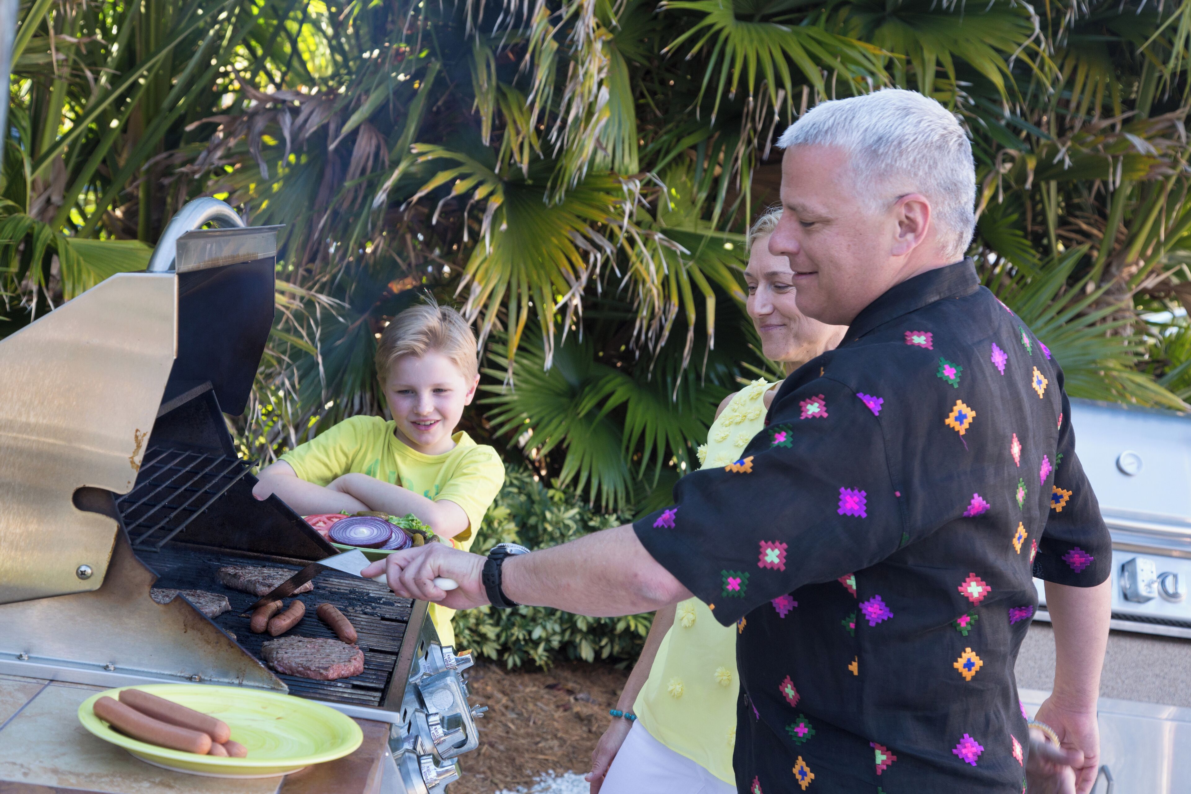 A man grilling hot dogs on an outdoor grill with a child and a woman nearby.