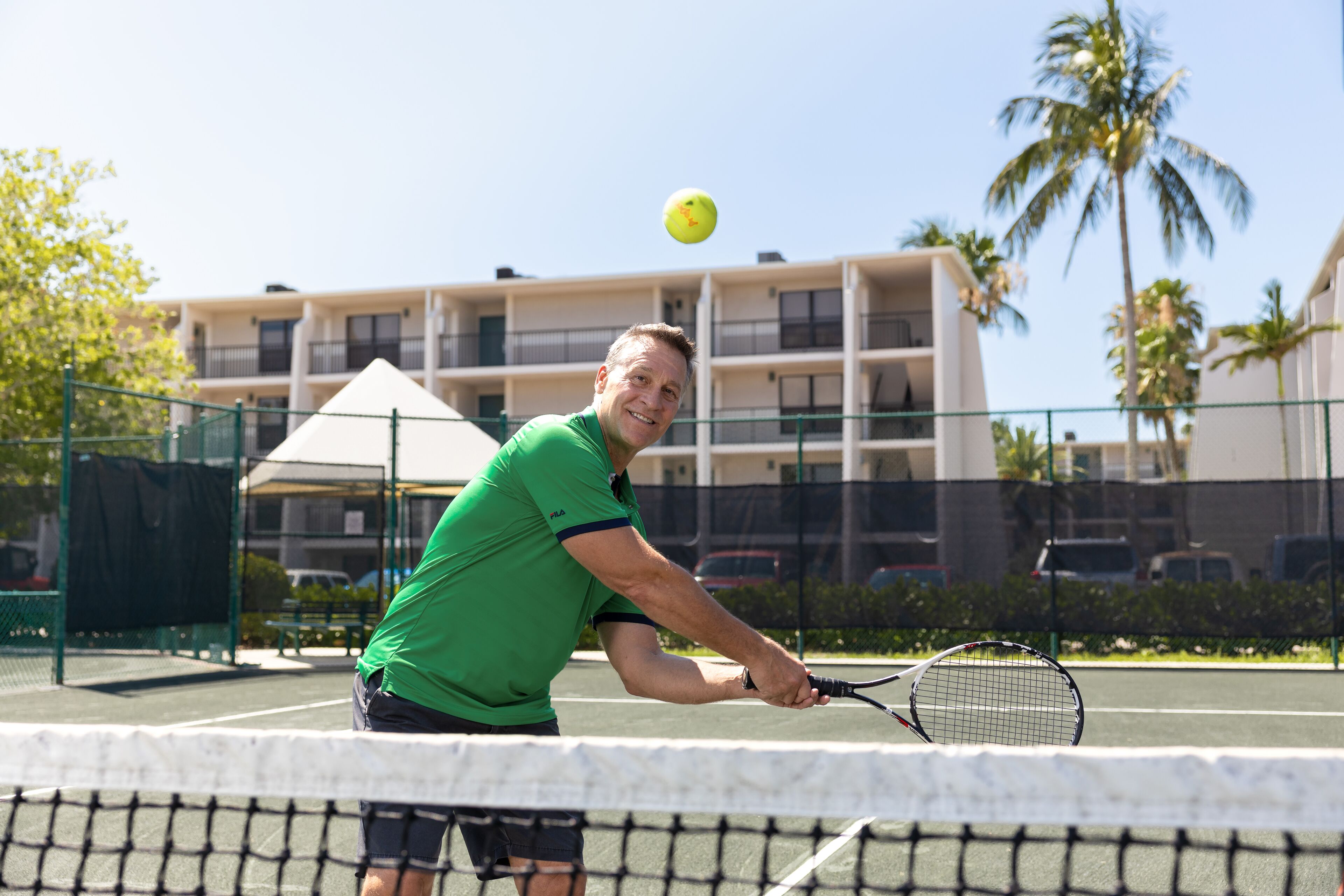 Ein Tennisplatz mit Netz, ein Spieler in einem grünen T-Shirt und ein Gebäude im Hintergrund.