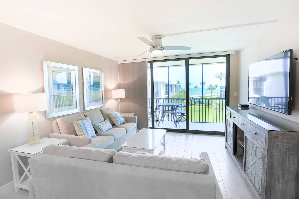 Ein modernes Wohnzimmer mit Sofa, Couchtisch und Fernseher. Es gibt einen Balkon mit Blick auf den Ozean und Palmen.