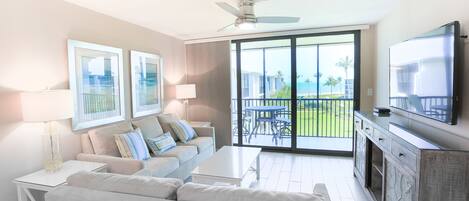 A modern living room with a sofa, coffee table, and TV. There is a balcony with a view of the ocean and palm trees.