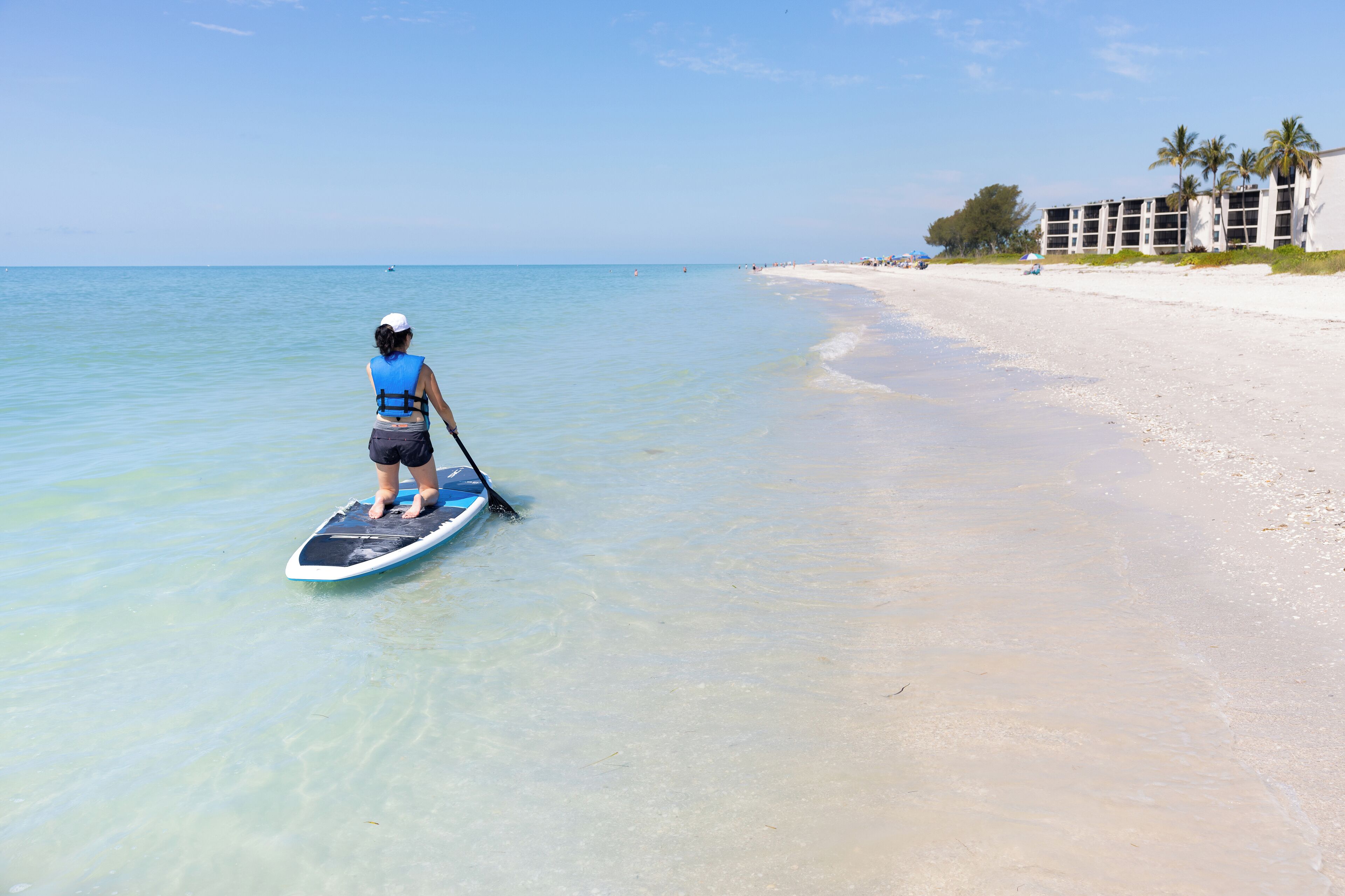 A person paddleboarding on clear shallow waters near a sandy beach with a building in the background.