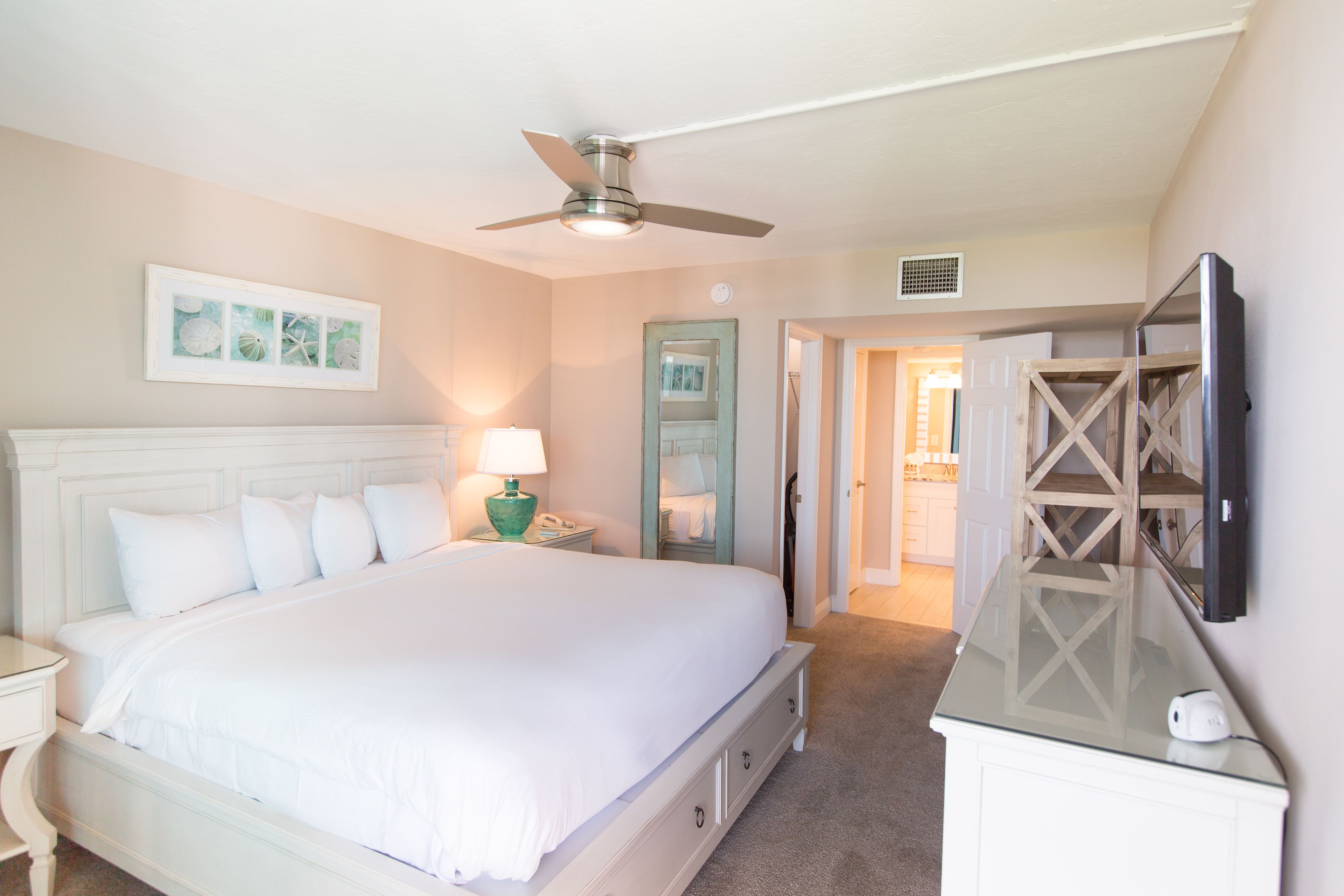 A bedroom with a large bed, a ceiling fan, a TV mounted on the wall, and a door leading to another room.