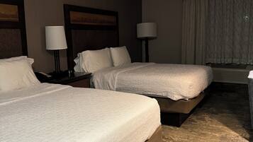 A hotel room with two beds, a nightstand with a lamp, a window with curtains, and a ceiling light.