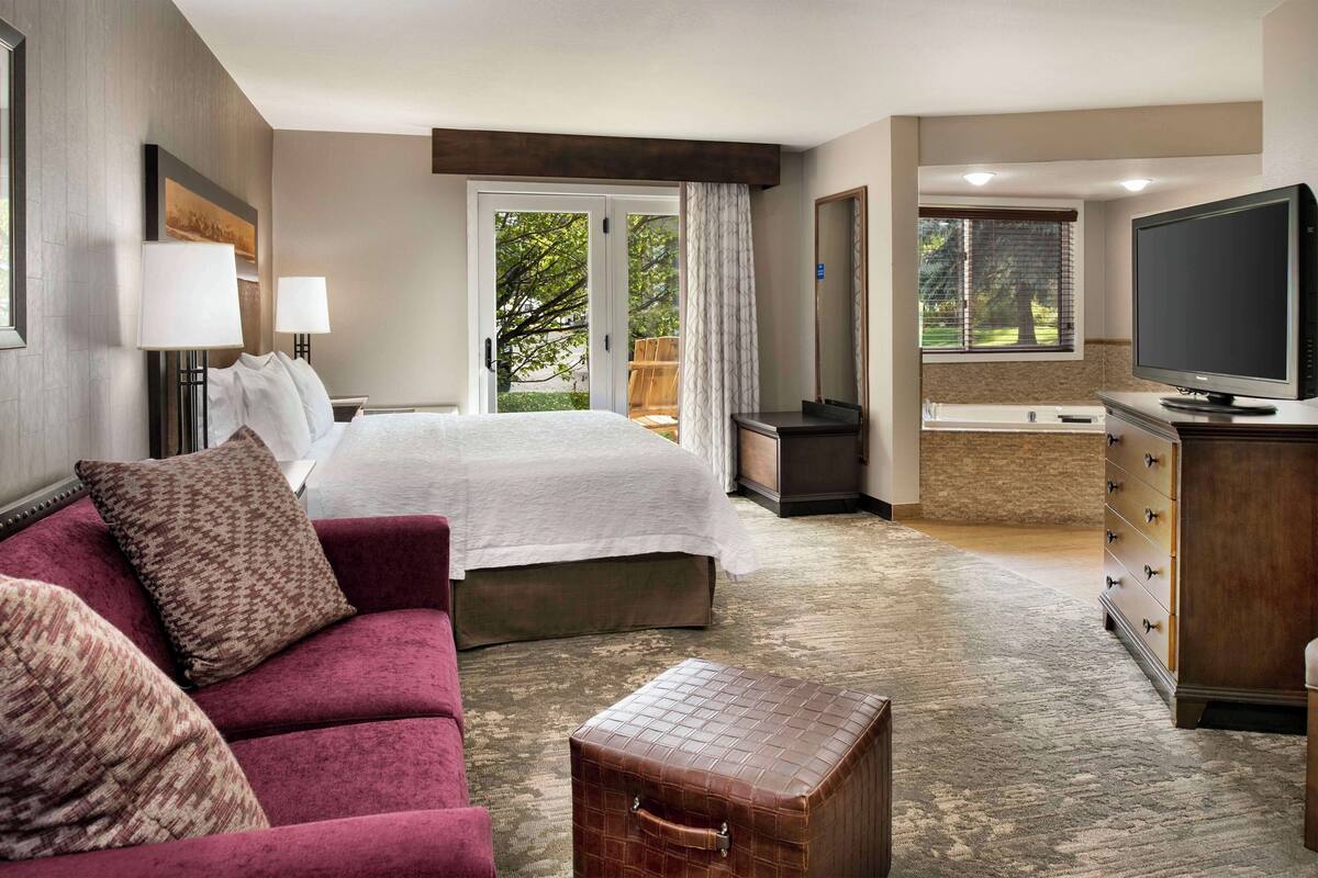 Quarto de hotel com cama, sofá, televisão, cômoda e mala.