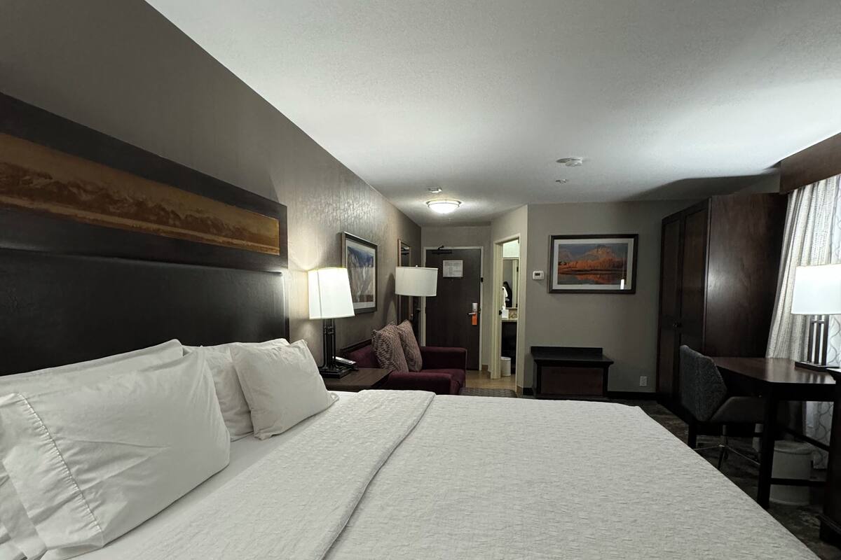 Quarto de hotel com uma cama grande, uma escrivaninha, uma cadeira, um abajur e um quadro na parede.