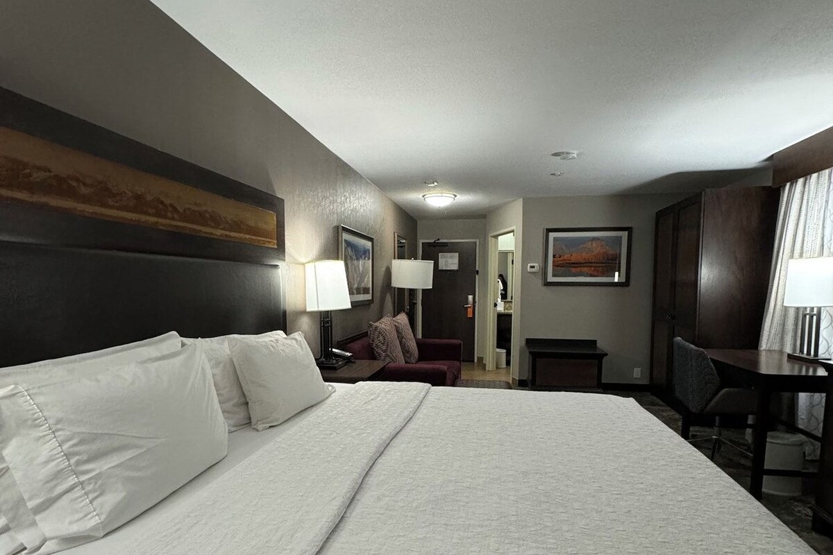 Een hotelkamer met een groot bed, een bureau, een stoel en uitzicht op de gang.